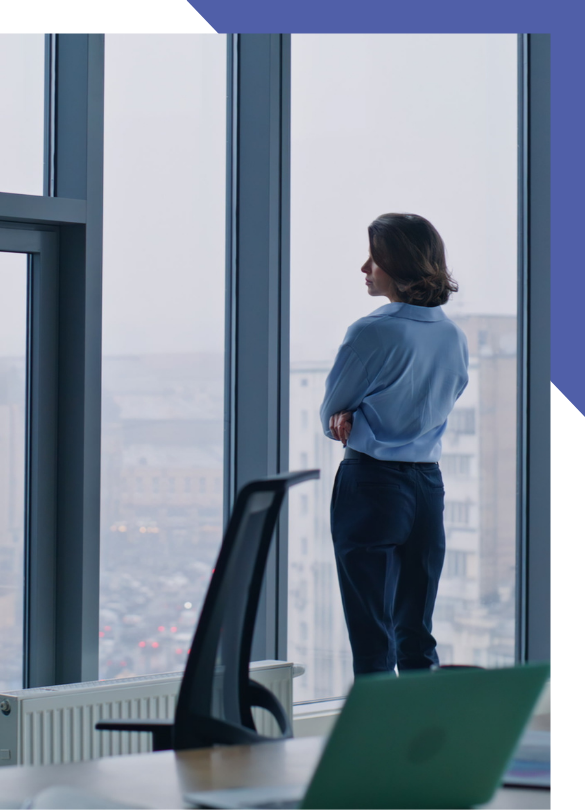 Woman Standing Alone Looking Out A High-rise Office Window With Arms Crossed, Representing The Isolation Of Leading Without Support