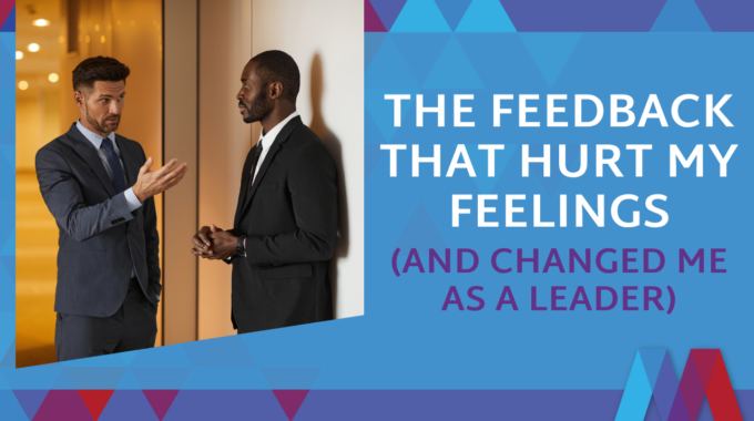 Feature Image For The Blog 'The Feedback That Hurt My Feelings (And Changed Me As A Leader)' Showing Two Male Colleagues In An Office Hallway, One Giving Feedback With An Open Hand Gesture.