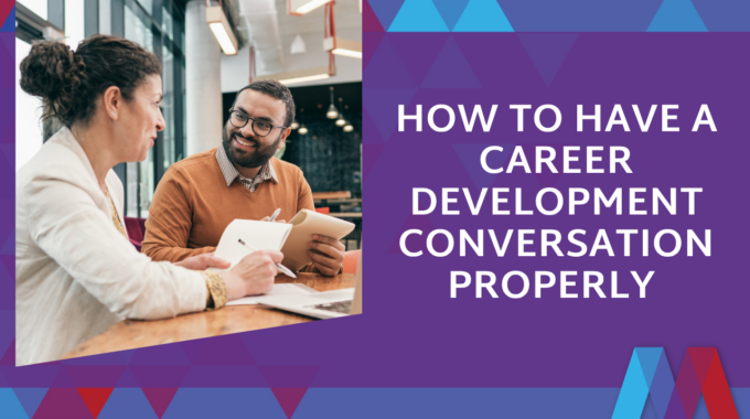 A Manager Takes Notes While Talking With A Smiling Team Member Across A Table In A Bright Office, Next To The Blog Title "How To Have A Career Development Conversation Properly" On A Purple Mosaic PD Background.